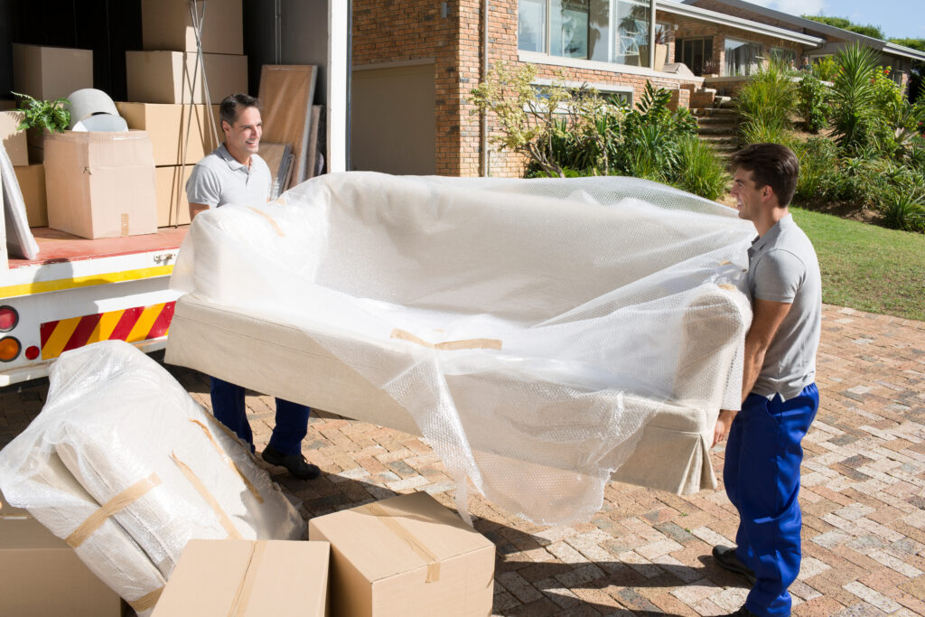 Two removal workers carrying a wrapped sofa from a house towards a moving van.